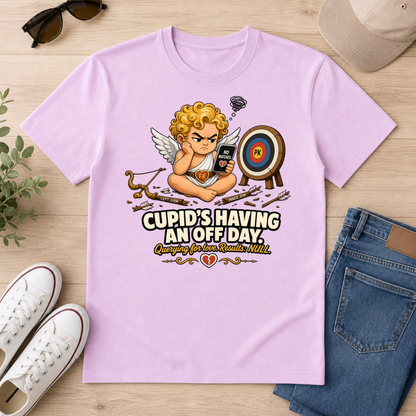 A light purple graphic t-shirt laid flat on a light wood background, surrounded by lifestyle props including blue jeans, white sneakers, sunglasses, and a beige baseball cap. The t-shirt features a cartoon cupid sitting cross-legged holding a phone that shows a storm cloud, with an arrow in his bow, a target, and the text 'CUPID'S HAVING AN OFF DAY' and 'Checking for Love, Straight Male'.