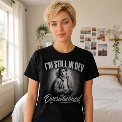 A woman with short blonde hair stands in a bedroom wearing a black graphic T-shirt with the text 'I'M STILL IN DEV Overwhelmed' and a grayscale portrait print on the front.