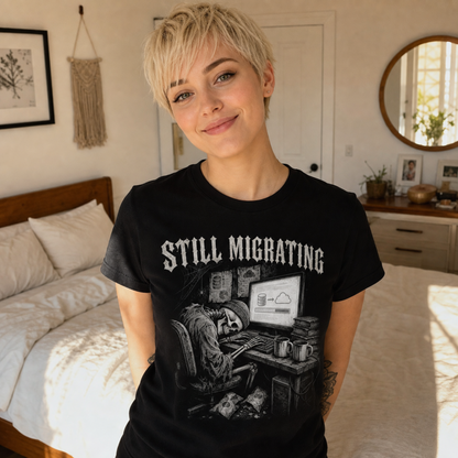 A woman with short blonde hair wearing a black graphic t-shirt that reads 'STILL MIGRATING' with a grayscale illustration of a person at a desk, standing in a bedroom.